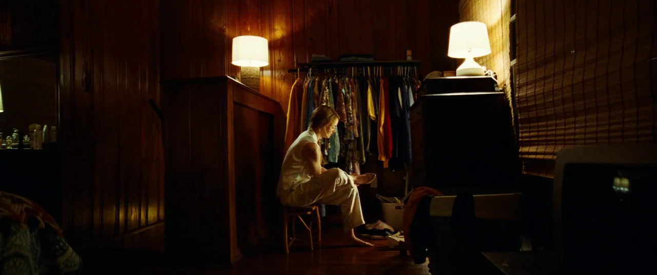 Kinds of Kindness (2024) movie still: medium — A dimly lit room with wood-paneled walls features a rack of colorful hanging clothes. A woman sits on…