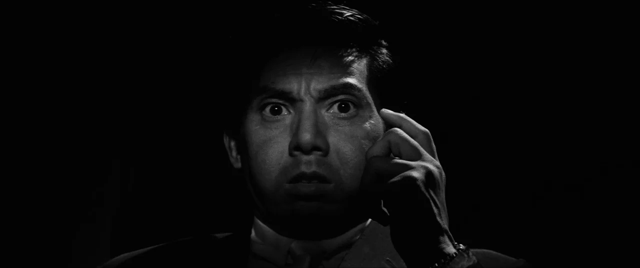 Branded To Kill (1967) movie still: closeup — A dark environment surrounds a man shown from the chest up. He stares ahead with wide eyes while…