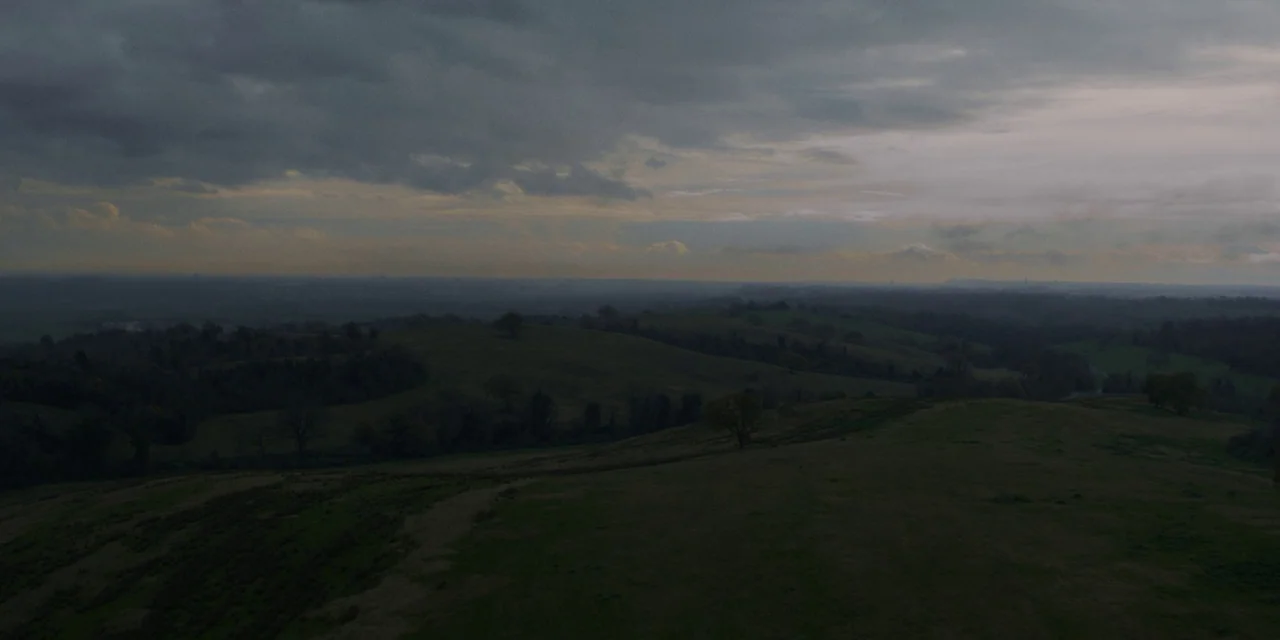 Immaculate (2024) movie still: establishing — An expansive view of dark rolling hills stretches toward a hazy distant horizon. A heavily clouded sky…