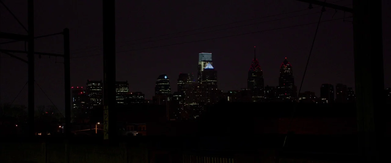 Creed (2015) movie still: establishing — A distant view of a city skyline at night under a dark sky. Several tall buildings feature illuminated…