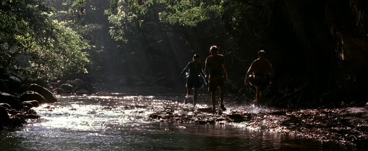 The Beach (2000) movie still: wide — A group of three people walks through a narrow shallow river in a forest. Sunlight breaks through the thick…