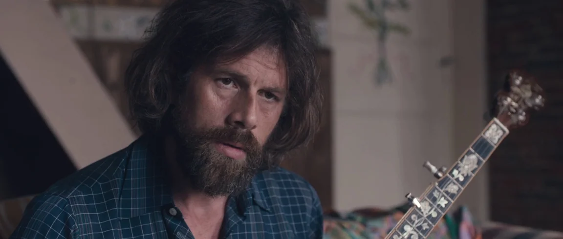 Broken Circle Breakdown (2012) movie still: closeup — A domestic space with decorative floral wall art visible in the background. A man with a thick…