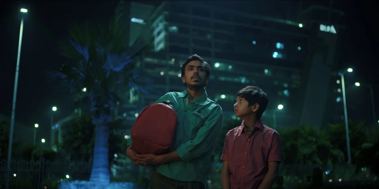 The White Tiger (2021) movie still: medium — A man and a boy stand at night in front of a modern corporate building. The man carries a red duffel…