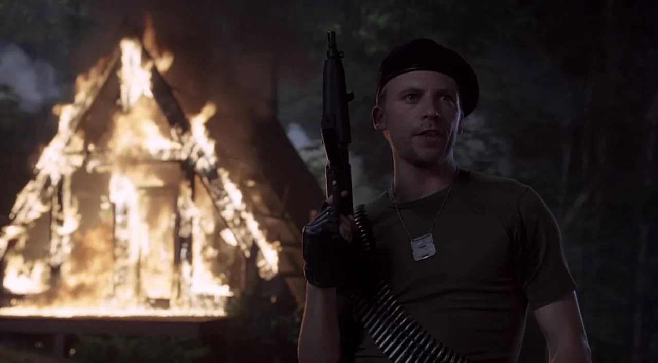 eXistenZ (1999) movie still: medium — A man in tactical gear stands before a burning wooden structure at night. Flames engulf the timber frame while…