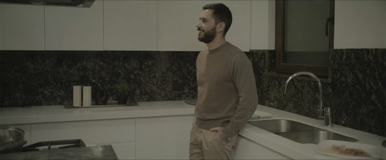 Los habitantes (2023) movie still: medium — A modern kitchen features white cabinetry and dark granite countertops with a stainless sink. A man in a…