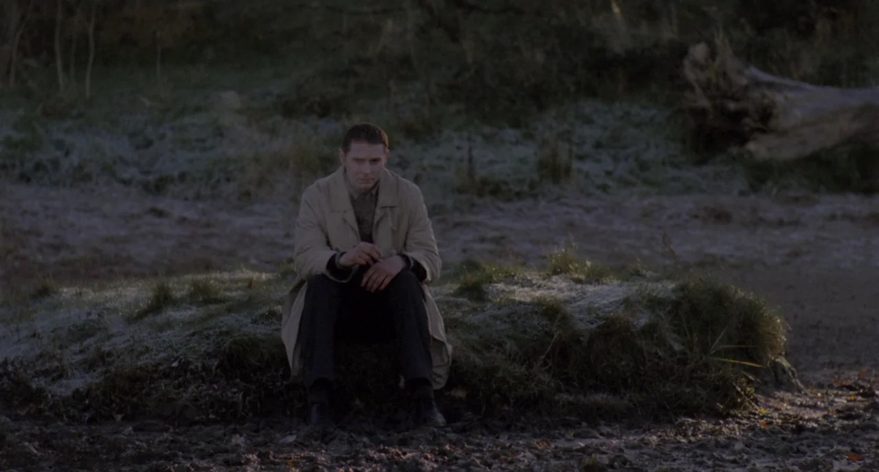 Possum (2018) movie still: medium — A man sits alone on a frosted, raised patch of earth in a muddy landscape. The surrounding terrain is barren and…
