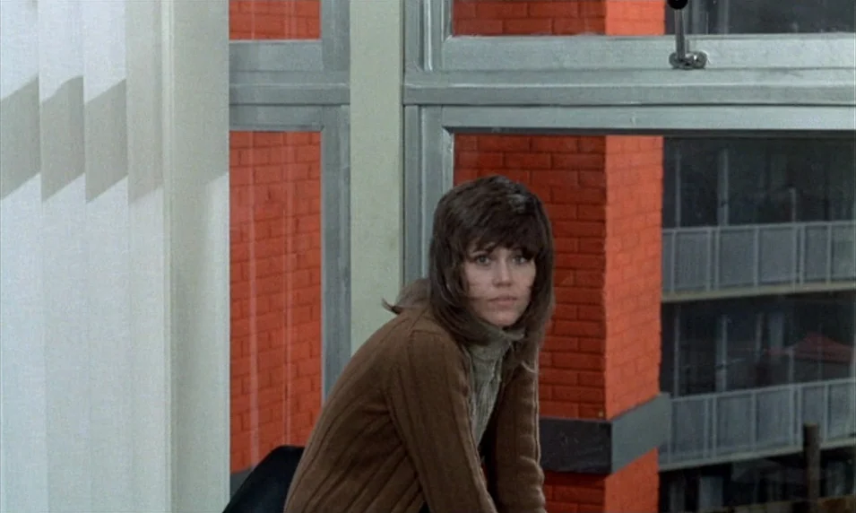 Tout Va Bien (1972) movie still: medium — A woman stands indoors in front of a window overlooking a brick building facade. She looks toward the…
