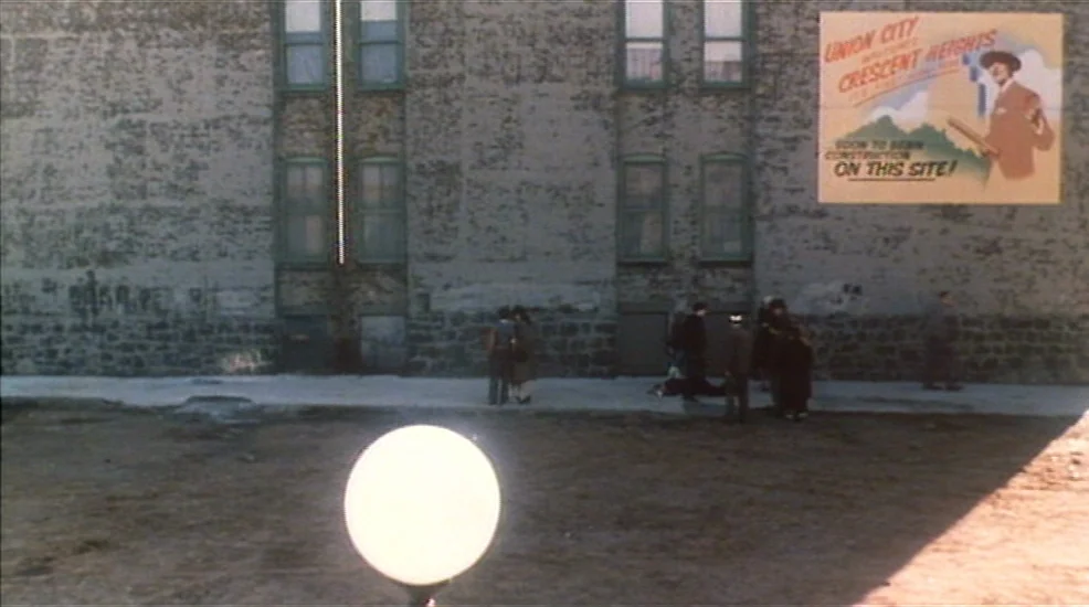 Union City (1980) movie still: wide — A vacant lot sits before a large apartment building with a promotional construction poster. Several people…