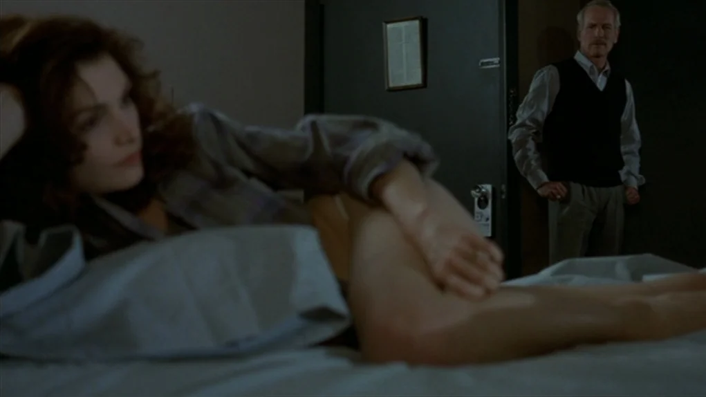 The Colour of Money (1986) movie still: medium — A muted hotel room contains a bed in the foreground and a doorway behind. A woman lies resting on…