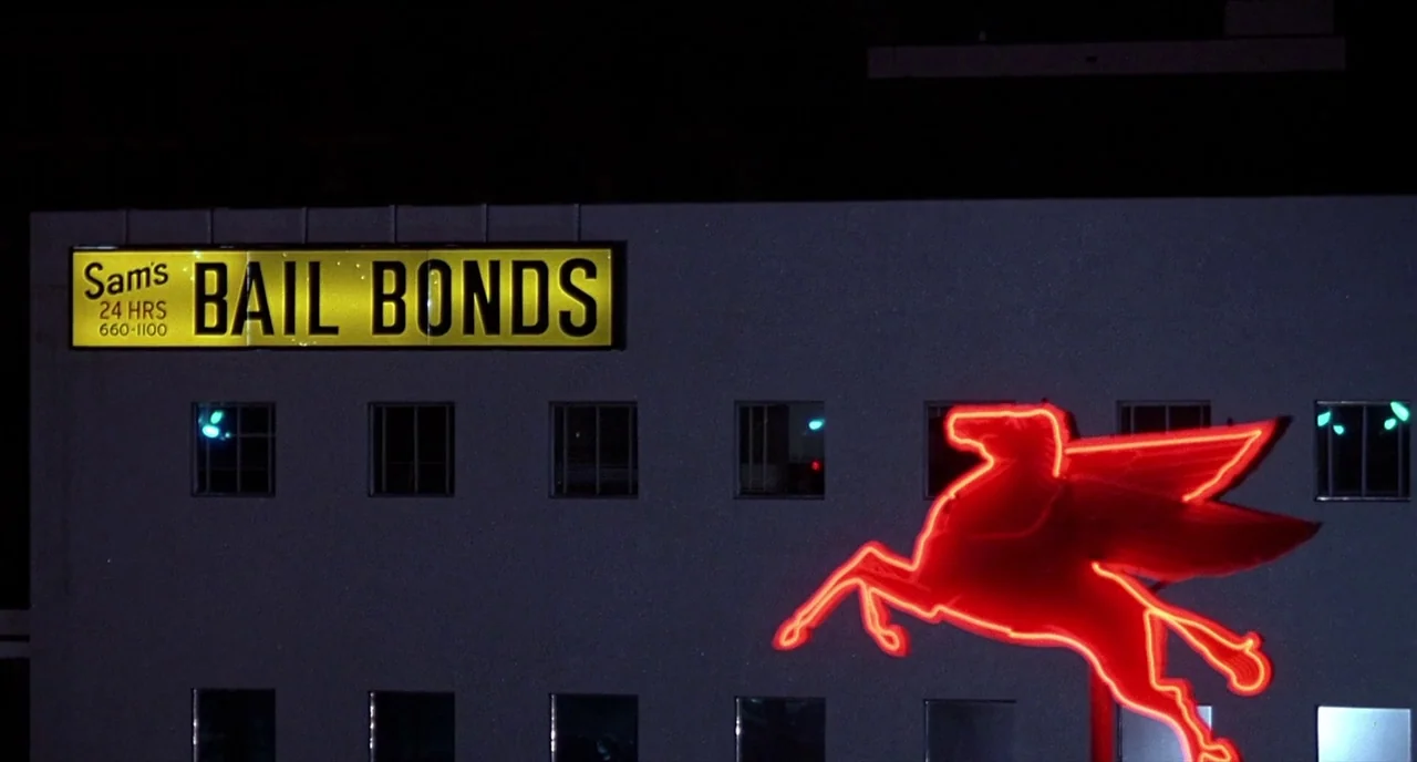 Barfly (1987) movie still: establishing — The exterior of a multi-story commercial building at night displays prominent signage. A glowing yellow…