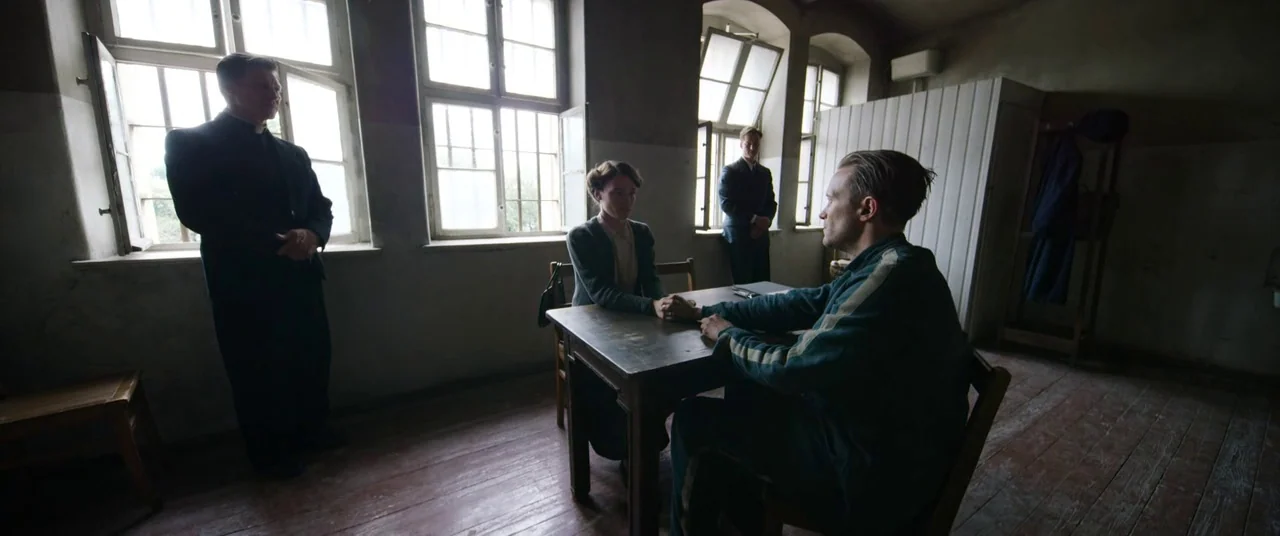 A Hidden Life (2019) movie still: wide — A stark 1940s visitation room with large multi-paned windows and bare, peeling plaster walls. A man and…