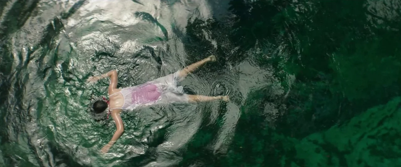 The Mad Hatter (2021) movie still: wide — A body floats on the surface of deep, dark green water. The subject is partially submerged in the center…