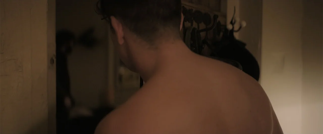 Bring Them Down (2025) movie still: closeup — A dimly lit residential hallway features a coat rack mounted on the wall. A shirtless man stands in…