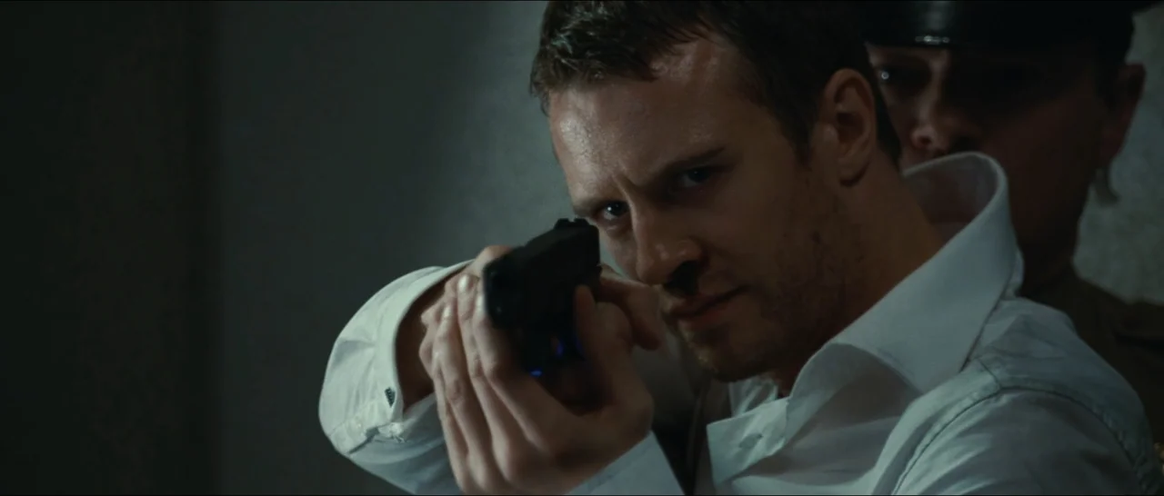 Slackers (2002) movie still: closeup — An interior space with a dimly lit wall behind the subjects. A man points a handgun forward with a second…