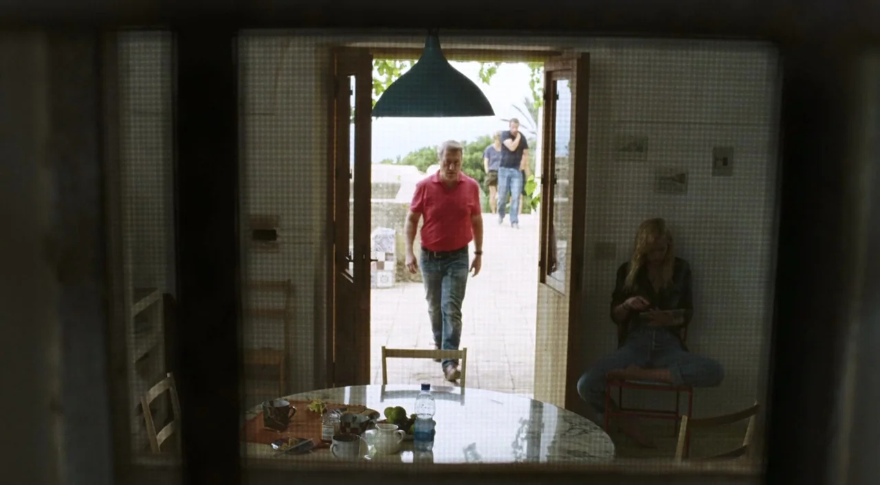 A Bigger Splash (2015) movie still: wide — An interior space viewed through a screen door looking out onto a bright terrace. A man walks through the…