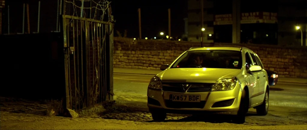 Kill List movie still: wide — A silver station wagon is parked on a paved lot beside a metal gate. A stone wall runs along the background under the…