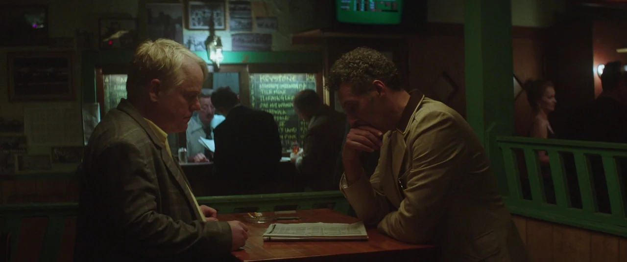 God’s Pocket (2014) movie still: medium — A dimly lit tavern interior features dark wood panelling and a green painted wooden partition. Two men sit…