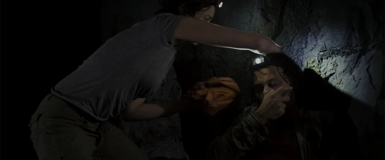 Black Water: Abyss (2020) movie still: medium — A dark, cramped stone cave interior with rugged, textured rock walls. A woman in a grey t-shirt…