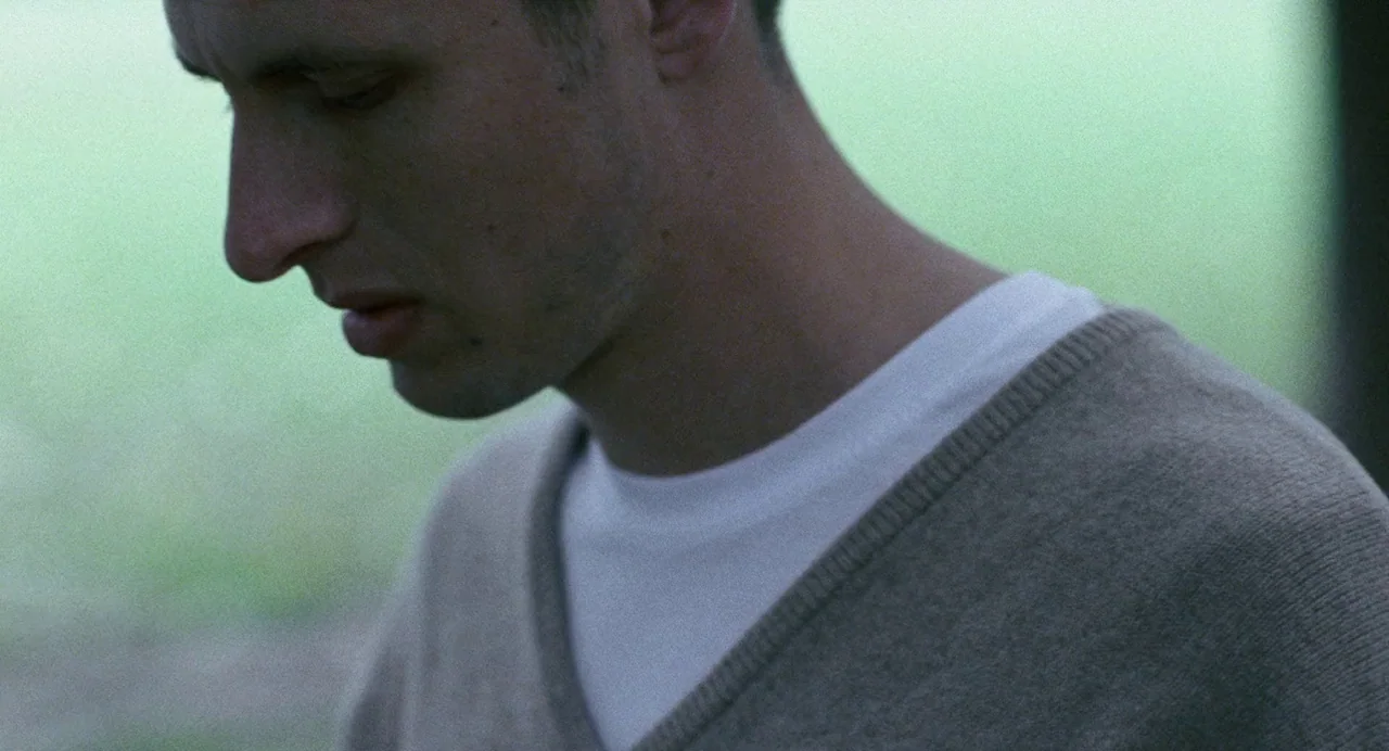Reprise (2006) movie still: closeup — A man stands outdoors in a soft-focused setting with greenery in the background. The man looks downward while…