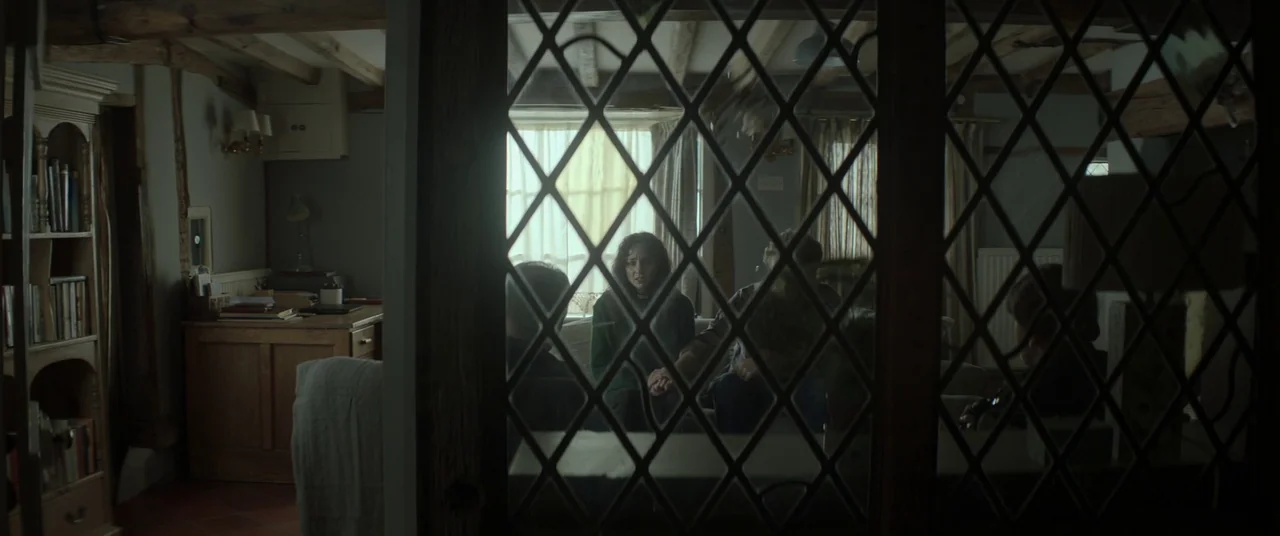 Lord of Misrule (2023) movie still: medium — A rustic interior with exposed wooden ceiling beams and a leaded glass room divider. People sit on a…