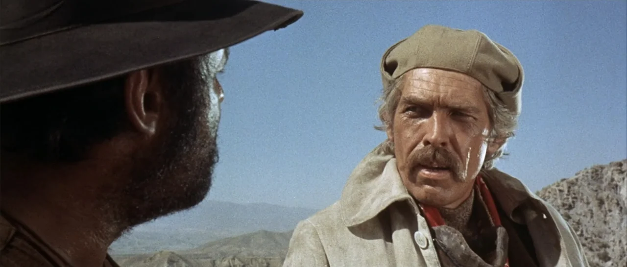 Duck, You Sucker (1971) movie still: medium — A dry, rocky desert landscape under a clear, bright blue sky. A man in a canvas jacket talks with…