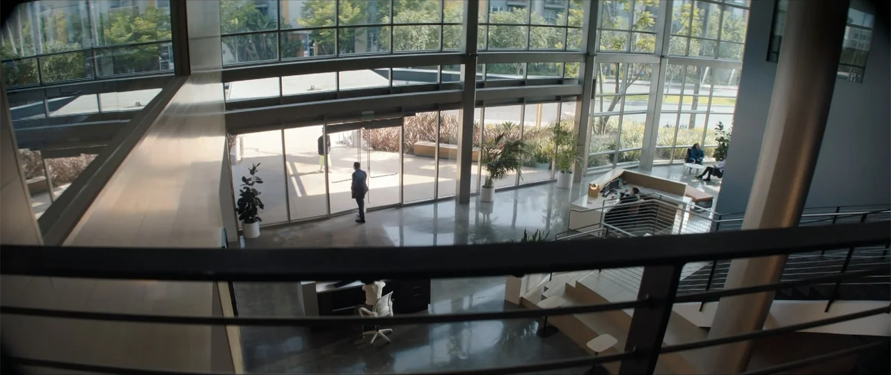 One of Them Days (2025) movie still: wide — A multi-level office lobby with high ceilings and floor-to-ceiling glass wall facade. A man stands at…