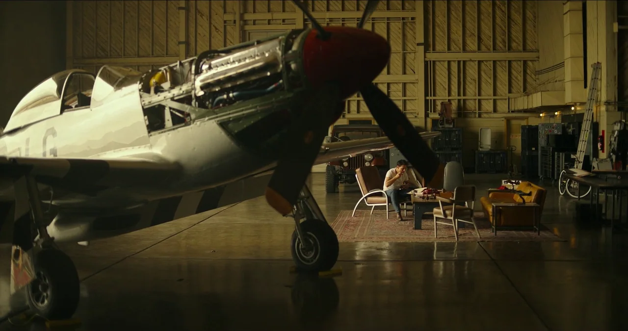 Top Gun: Maverick (2022) movie still: wide — A man sits in an armchair inside a large, industrial airplane hangar. A vintage propeller plane is…
