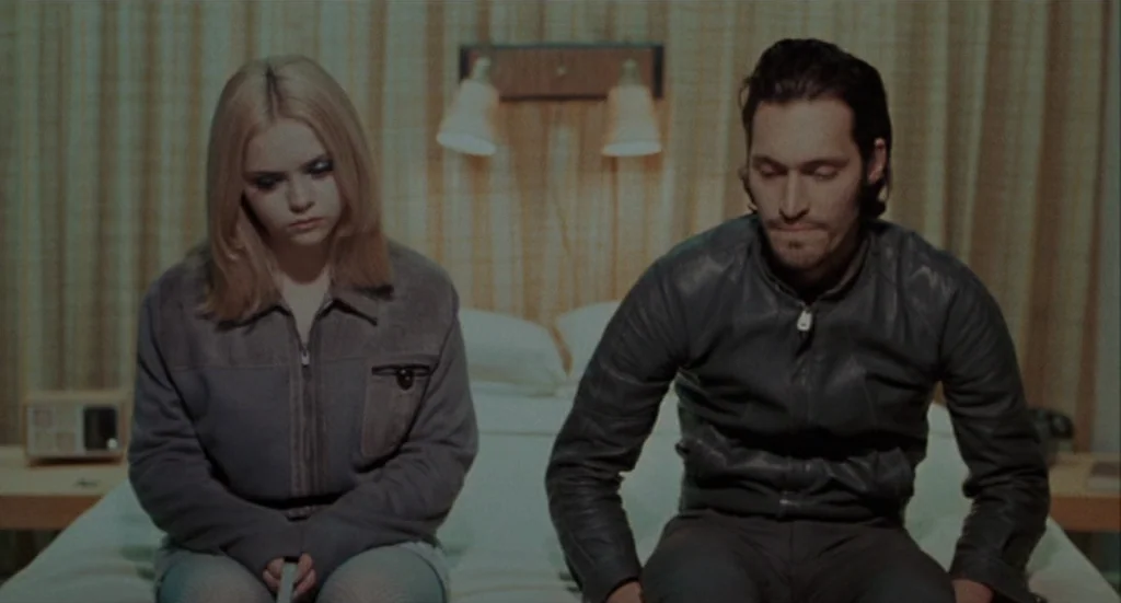 Buffalo ’66 (1998) movie still: medium — A motel room with striped textured wallpaper and bedside lamps mounted on the wall. A man and a woman sit…
