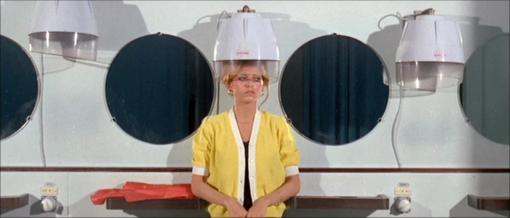 Made in U.S.A. (1966) movie still: medium — A vintage hair salon features white walls with circular mirrors and rows of hood dryers. A woman in a…