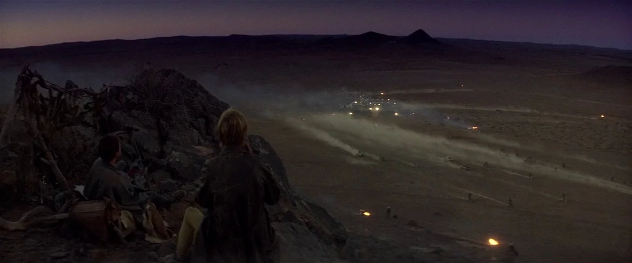 Mad Max 2 (1981) movie still: wide — A vast, dusty desert expanse at dusk with a line of moving vehicle lights. Two figures sit on a rocky…