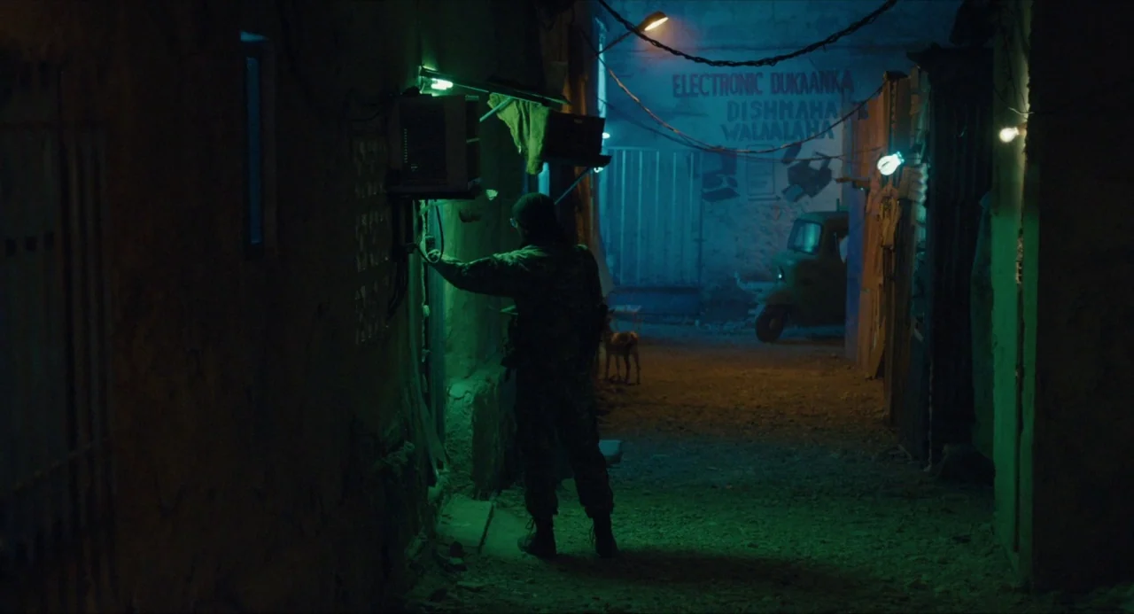 Submergence movie still: wide — A narrow dirt pathway is lined by dark walls with hanging electrical cables. A person stands at a wall inspecting a…