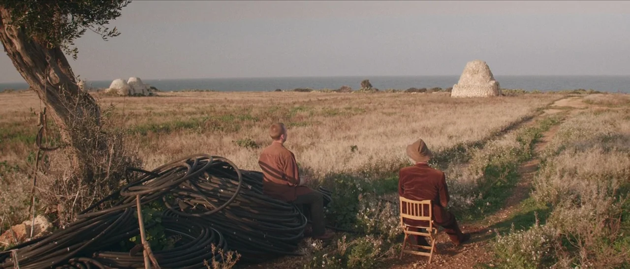 Spring (2014) movie still: wide — A dry coastal field features a large gnarled tree and coiled black irrigation pipes. Two men sit in the open…