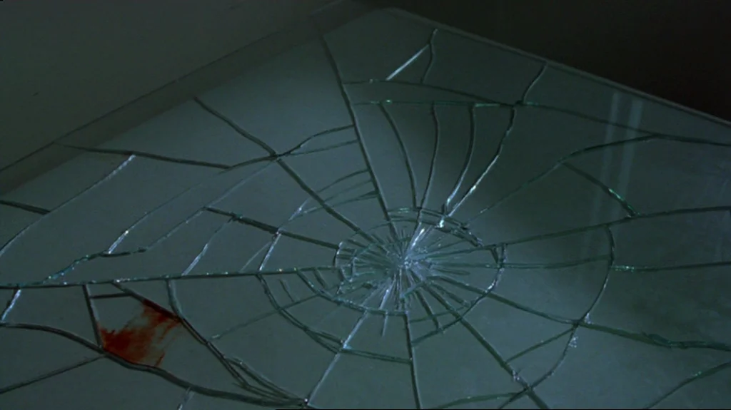 Spider (2002) movie still: closeup — A flat glass surface displays a complex spiderweb pattern of cracks and fissures. A singular red bloodstain…