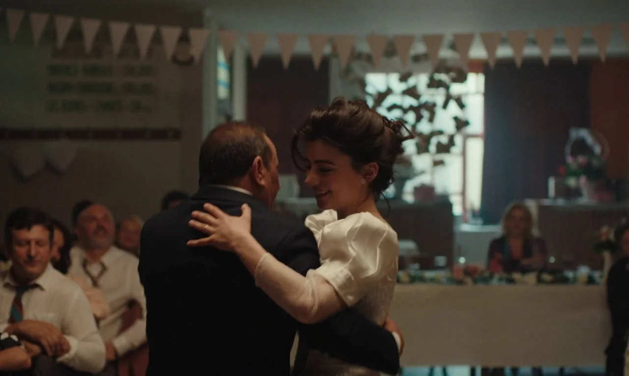 Maldoror (2024) movie still: medium — An indoor event space decorated with paper pennants hanging from the ceiling. A bride and an older man are…
