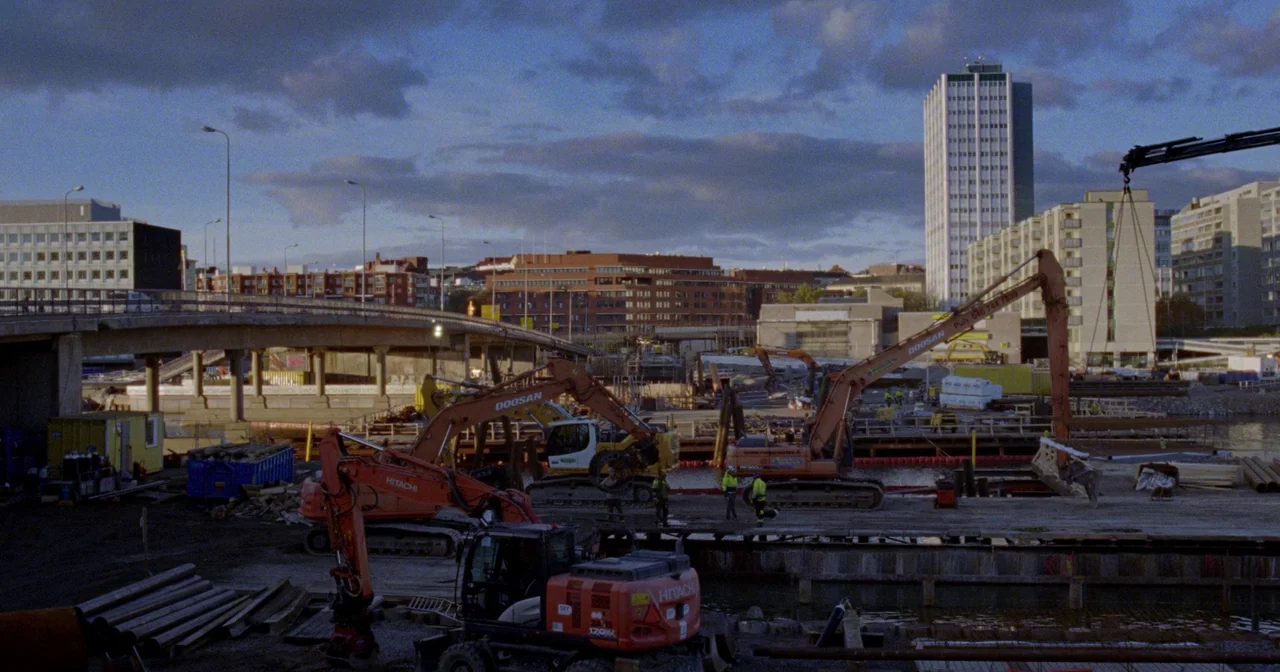 Fallen Leaves (2023) movie still: wide — An industrial construction site sits beside a river under a cloudy late-afternoon sky. Several orange…