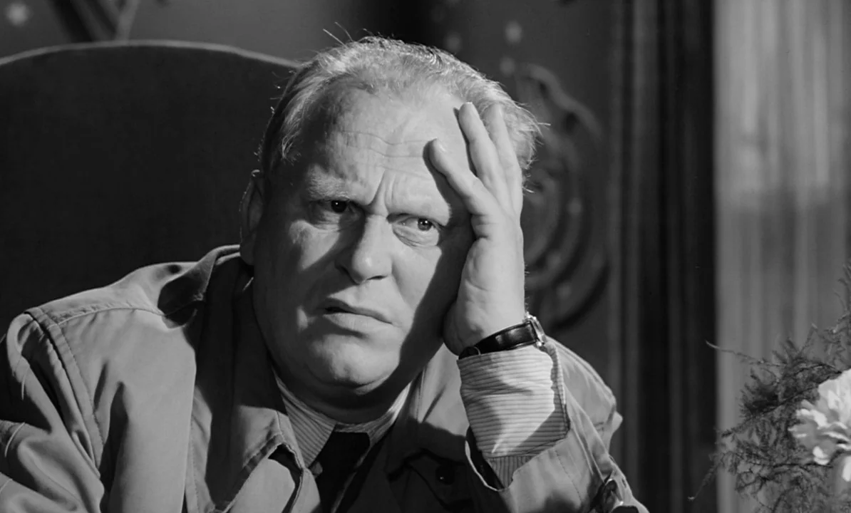 The 1,000 Eyes of Dr. Mabuse (1960) movie still: closeup — A man sits at a desk in a dark room with patterned wall decorations. He holds his head…