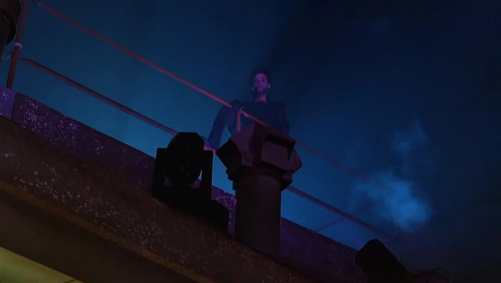 Graffiti Bridge (1990) movie still: wide — A dark urban rooftop at night with metal railings and industrial ventilation ducts. A man sits perched on…