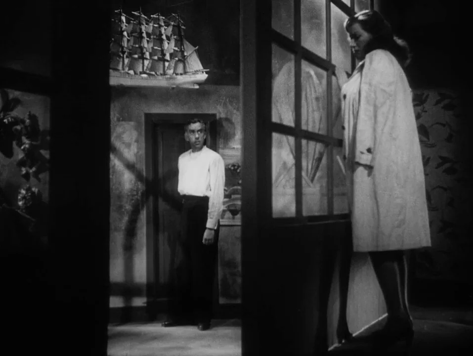 A Ship Bound for India (1947) movie still: medium — A dark apartment room features a glass partition and a prominent ship model. A woman in a trench…