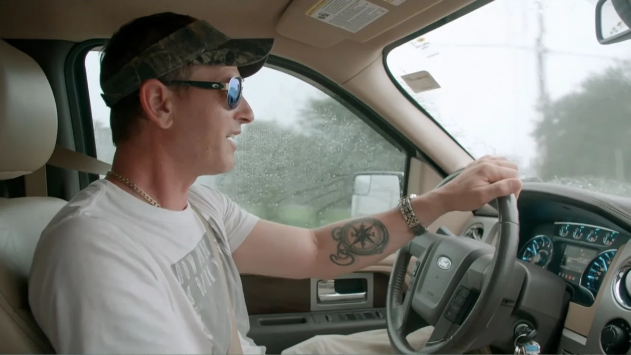 Mommy Dead and Dearest (2017) movie still: medium — The interior of a modern Ford truck during a light rainstorm. A man wearing a camo visor and…