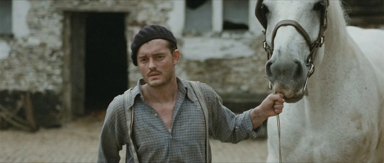 Suite Française (2015) movie still: medium — A rustic farmyard featuring an old stone wall background and a dirt ground surface. A man in a beret…