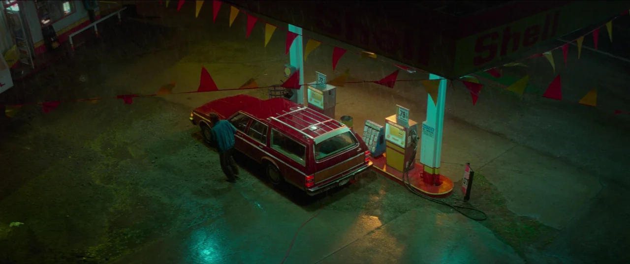White Noise (2022) movie still: wide — A rain-slicked Shell gas station at night features colorful plastic flags under a canopy. A lone man stands…