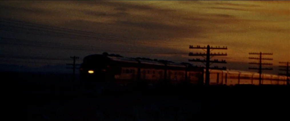 The Getaway (1972) movie still: wide — A passenger train travels horizontally across a vast, dark landscape during twilight hours. Silhouetted…