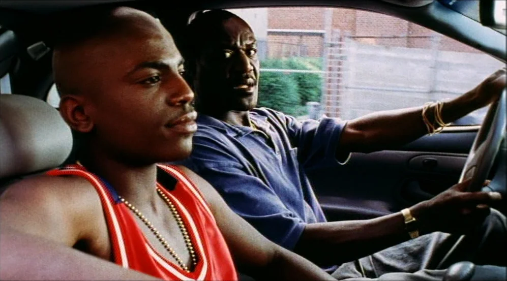 Clockers (1995) movie still: medium — Two men sit inside a vehicle during the day with urban surroundings visible outside. The driver steers while…