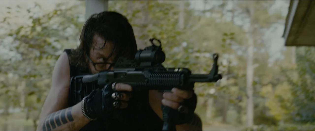 Ambush (2020) movie still: medium — A man stands outdoors near trees while holding a firearm. He aims a black rifle with a red dot sight.