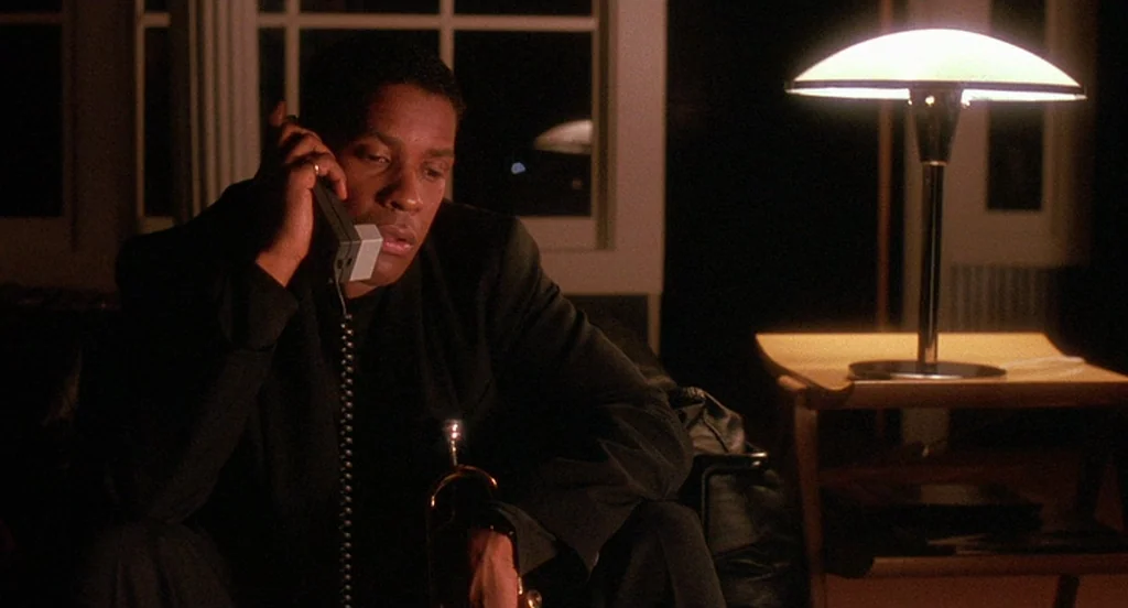 Mo’ Better Blues (1990) movie still: medium — A dimly lit interior with a dark window backdrop and a side table lamp. A man in a dark suit speaks on…