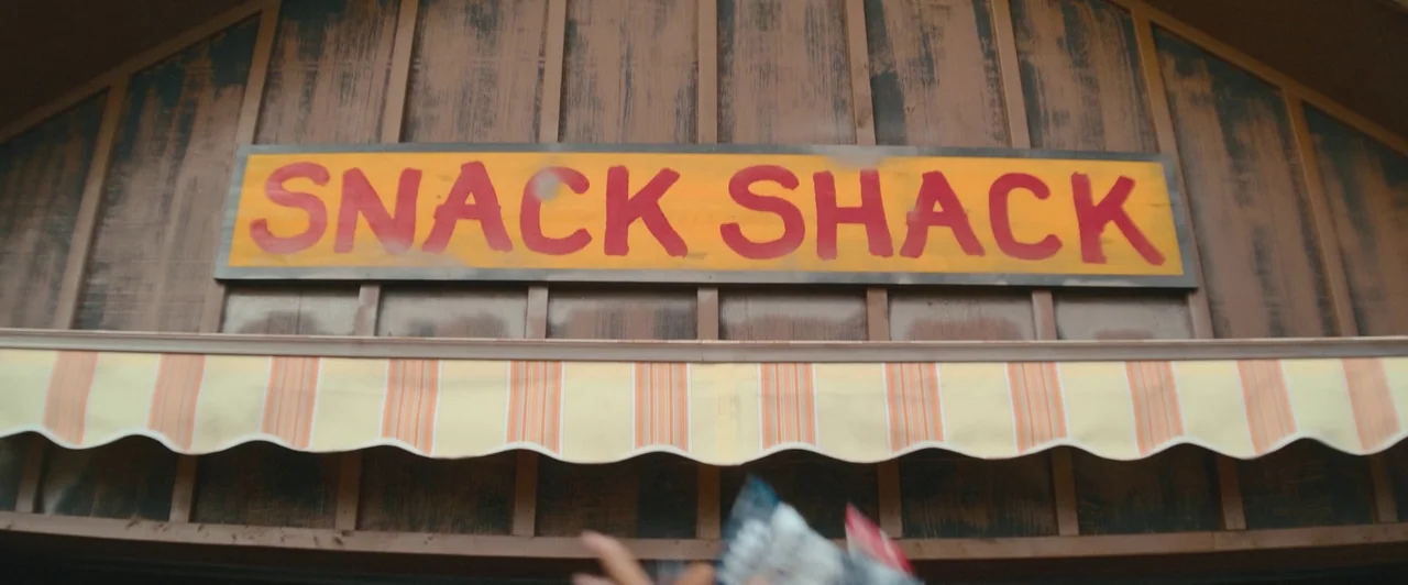 Snack Shack (2024) movie still: medium — A rustic wooden structure features a prominent hand-painted sign reading Snack Shack. A striped fabric…