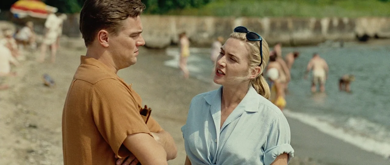 Revolutionary Road (2008) movie still: medium — A populated sandy beach features a sea wall and blurred figures in the background. A man and a woman…