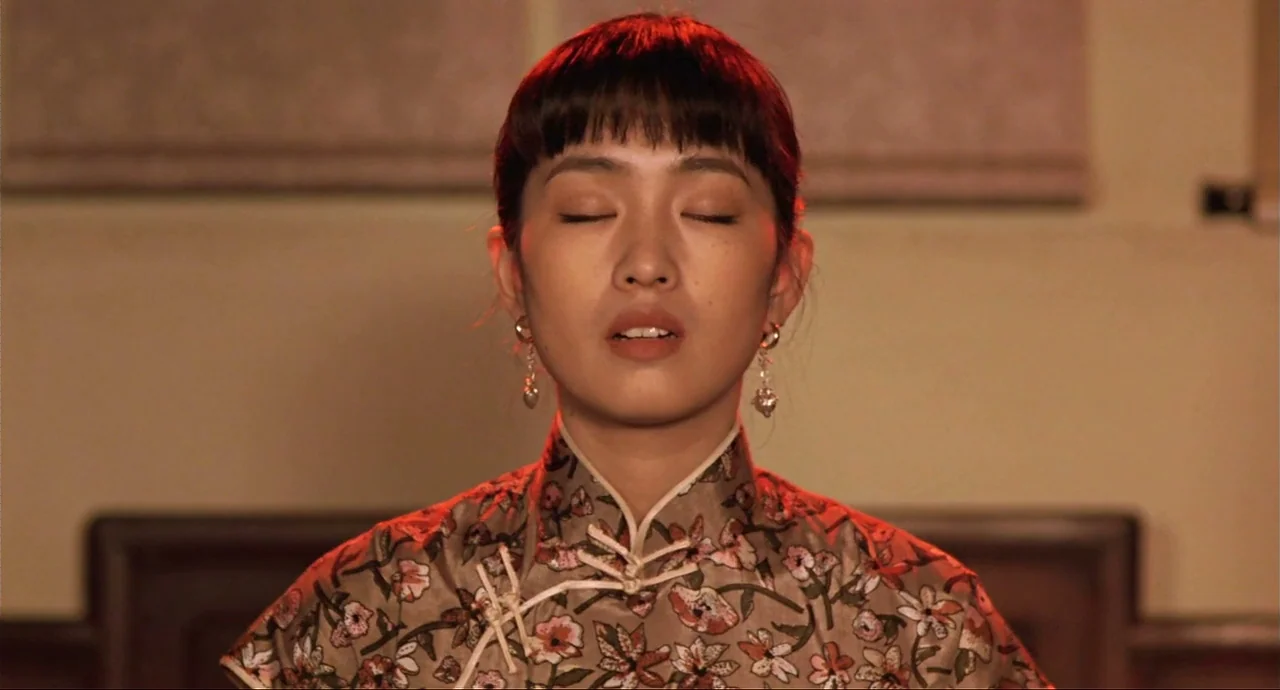 Raise The Red Lantern movie still: closeup — A woman stands in a dimly lit room with simple wall decor visible behind her. She keeps her eyes closed…