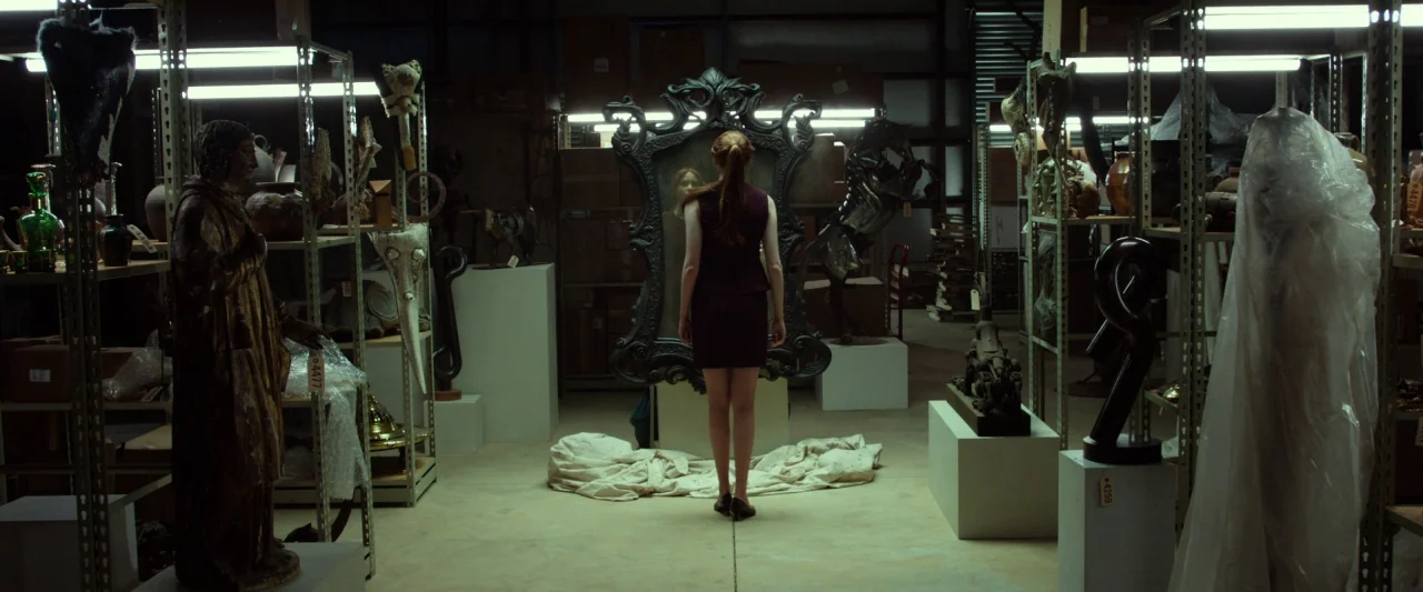 Oculus (2013) movie still: fullbody — A cavernous storage space features industrial shelving units filled with various art objects. A young woman…