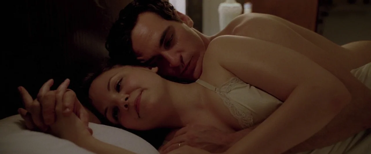 Walk The Line (2005) movie still: closeup — A man and woman lie together in bed beneath white linens. The two individuals are embracing while…